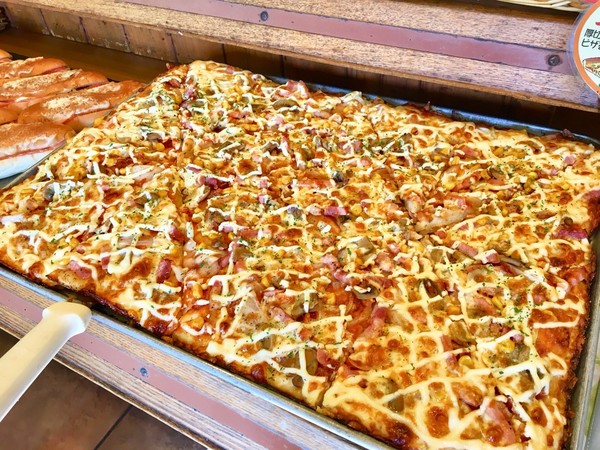 「鉄板ピザ」(１ピース248円)。たっぷりの具材とチーズをのせて焼き上げた大きなピザ。焼きたてはチーズがとろ～り！　ベーコンが厚切りになってより美味しく具沢山に。ピザが焼きあがると店内中にピザのいい匂いがあふれて、ついつい食べたくなってしまう一品。